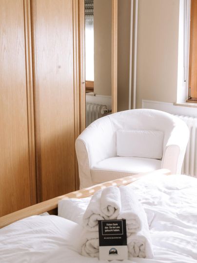 Een gezellige slaapkamer met een witte stoel en een houten kast. Vers gevouwen handdoeken liggen op het bed.