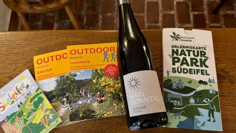 Een fles wijn staat op een tafel naast verschillende brochures over uitstapjes. De brochures tonen verschillende regio's en outdoor-activiteiten.