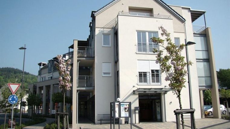 Ein modernes Gebäude mit klaren Linien und großen Fenstern. Vor dem Gebäude sind bepflanzte Flächen und eine kleine Straße sichtbar.