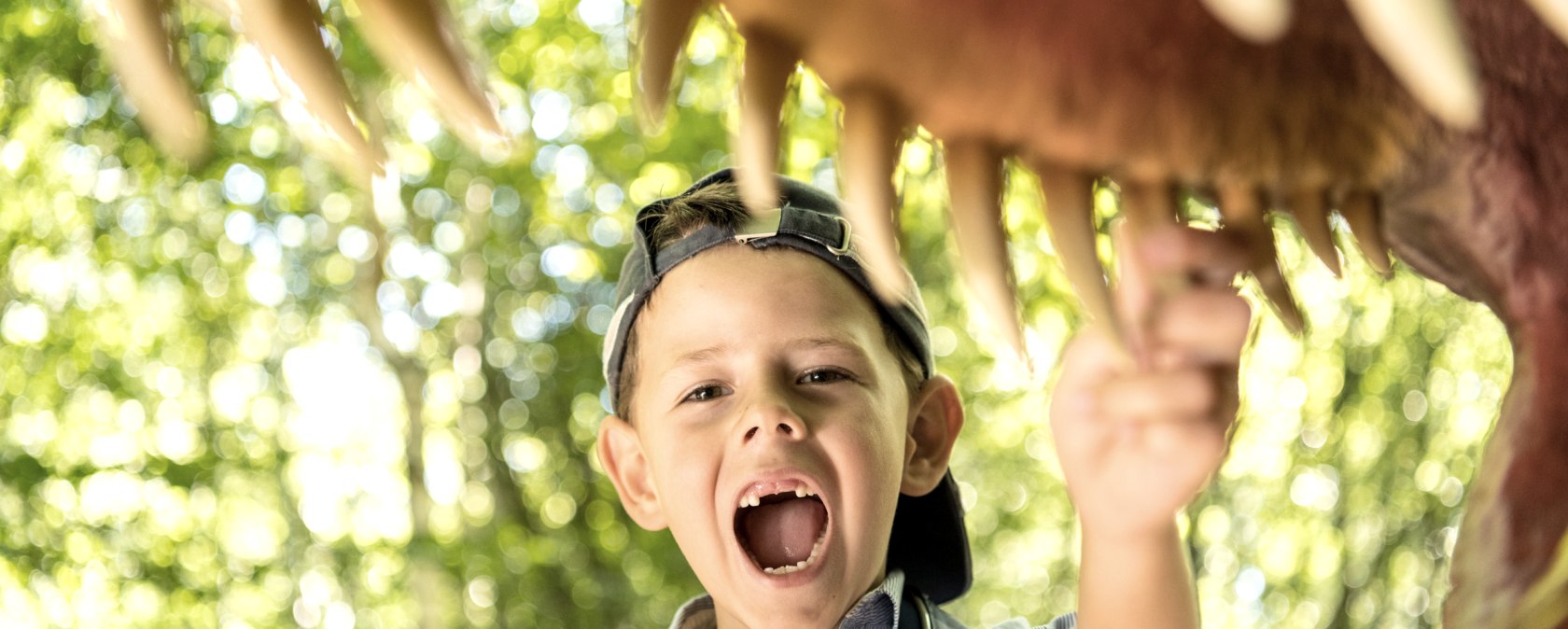 A boy under a dinosaur model with his mouth open, pointing to his teeth. In the background are trees.