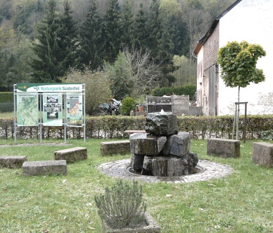 Ein Brunnen aus Steinen in Mettendorf, umgeben von B&auml;nken. Im Hintergrund eine Infotafel des Naturparks S&uuml;deifel und ein Geb&auml;ude., &copy; Felsenland S&uuml;deifel Tourismus, Christian Calonec-Rauchfuss