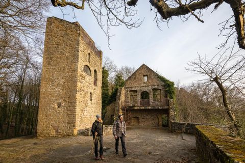 Zwei Wanderer stehen vor einer alten Ruine in einem Waldgebiet. Die Steinmauern des Gebäudes sind von Pflanzen überwuchert und umgeben von Bäumen.
