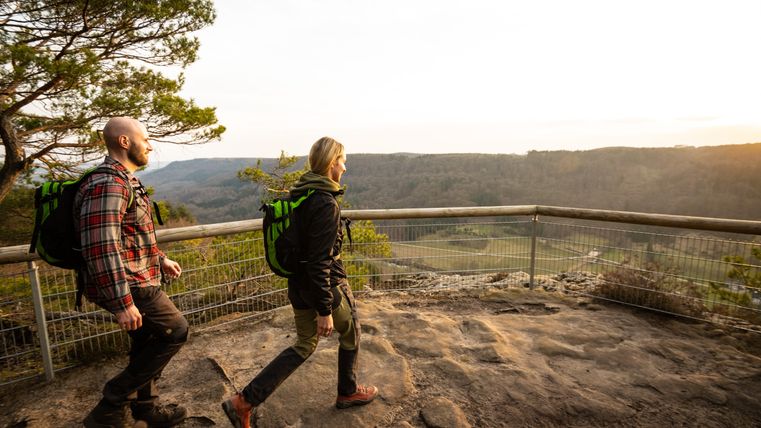 Zwei Wanderer auf einem felsigen Aussichtspunkt mit Blick auf eine bewaldete Landschaft bei Sonnenuntergang.