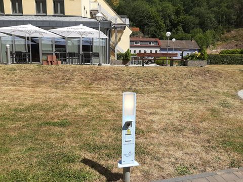 Ein heller, moderner Beleuchtungskörper steht auf einer Wiese vor einem Gebäude. Im Hintergrund sind Tischdecken und Sonnenschirme eines Außenbereichs sichtbar.