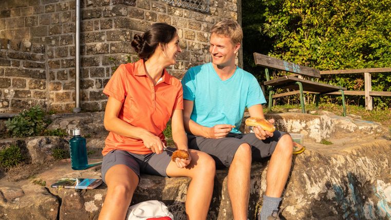 Ein Paar sitzt entspannt auf einer Treppe und lächelt sich an. Sie genießen die Natur und haben Freunde in der Nähe.