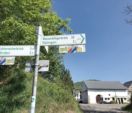 Wegweiser zeigt Entfernungen zu Echternacherbrück, Minden, Wasserbilligerbrück und Ralingen. Im Hintergrund ein Gebäude, blauer Himmel., © Tourist-Information Bitburger Land, Steffi Wagner