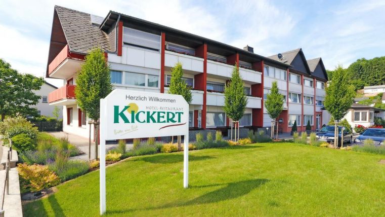 Ein modernes Gebäude mit einem einladenden Schild "Herzlich Willkommen" steht im Vordergrund. Der gepflegte Garten ist mit Bäumen und frischem Gras gestaltet.