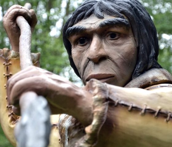 Pr&auml;historische Figur mit Speer im Dinopark, umgeben von B&auml;umen. Audiotour-Interface sichtbar.