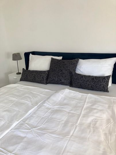 Ein modernes Schlafzimmer mit einem großen Bett, das mit weißen Laken und mehreren Kissen dekoriert ist. Dies wird von einem Nachttisch mit einer kleinen Lampe ergänzt.