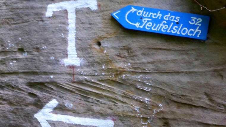 Ein Fels mit weißen Pfeilen und einem blauen Wegweiser. Der Wegweiser zeigt in Richtung "Teufelsloch".