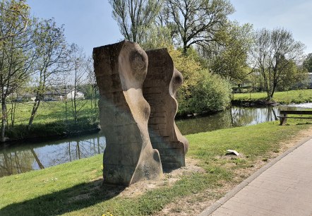 Stone sculpture on the riverbank, surrounded by green landscape and trees. A clear sky and a path are also visible., © TI Bitburger Land, Steffi Wagner