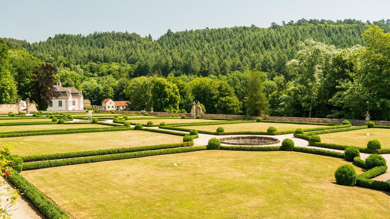 Ein gepflegter Garten mit symmetrischen Hecken und grünen Buchsbäumen. Im Hintergrund sind Wälder und ein helles Haus zu sehen.