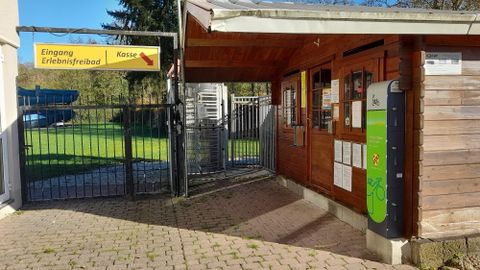 Eingang zum Erlebnisfreibad mit einem Holzgebäude und einem Zaun. Auf einem Schild steht "Eingang Erlebnisfreibad" und "Kasse".