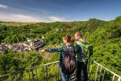 Ein Paar steht auf einem Aussichtspunkt und blickt auf eine malerische Stadt umgeben von grüner Landschaft. Der Himmel ist klar und die Natur wirkt einladend.