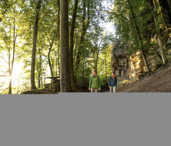 Zwei Wanderer gehen auf einem Waldweg entlang einer Felswand. Die Sonne scheint durch die B&auml;ume und taucht die Szene in warmes Licht., &copy; Dominik Ketz