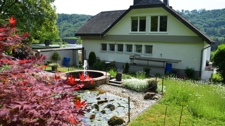 Ein schönes Haus mit einem gepflegten Garten und einem Teich. Im Hintergrund sind sanfte Hügel und Bäume zu sehen.