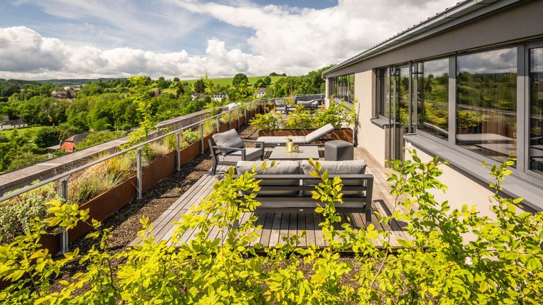 Eine moderne Dachterrasse mit bequemen Sitzgelegenheiten und üppiger Vegetation. Im Hintergrund ist eine grüne Landschaft und ein strahlend blauer Himmel zu sehen.