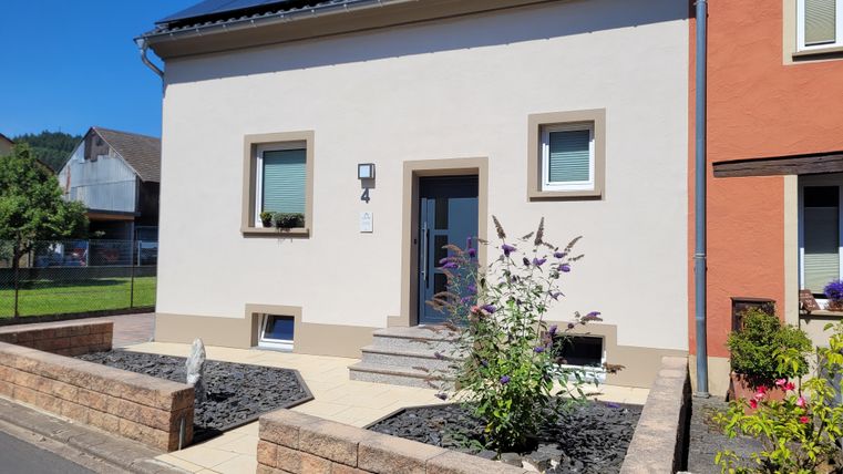 Ein modernes Haus mit einer hellen Fassade und einem kleinen Vorgarten. Der Eingang ist durch Stufen erreichbar und es gibt einige Pflanzen vor dem Haus.