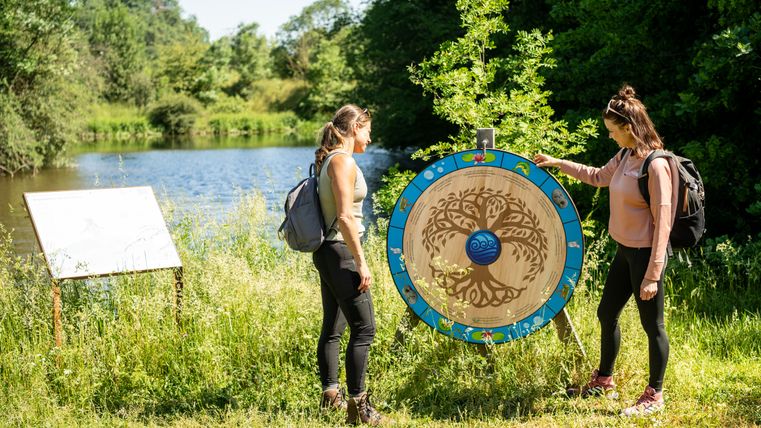 Zwei Frauen stehen an einem dekorativen Holzrad am Ufer eines Sees.