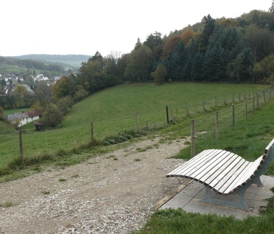 Een gebogen houten bank staat op een heuvel die uitkijkt over Mettendorf, omringd door groene weiden en bomen., © Felsenland Südeifel Tourismus GmbH, Christian Calonec-Rauchfuss