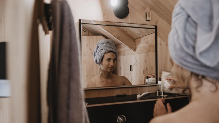 Eine Frau mit einem Handtuch auf dem Kopf schaut in den Spiegel. Der Raum ist hell und aus Holz gestaltet.
