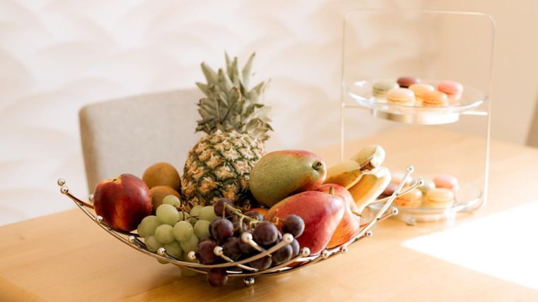 Een stijlvolle fruitschaal met appels, druiven, een ananas en een peer staat op een tafel. Achter de schaal staat een rek met macarons.