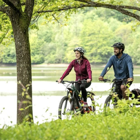 Zwei Radfahrer fahren lächelnd auf einem Weg am Stausee Bitburg entlang, umgeben von grüner Natur und Bäumen., © Eifel Tourismus GmbH, Dominik Ketz