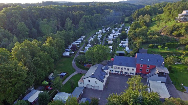 Ein malerischer Campingplatz umgeben von grünen Bäumen. Im Hintergrund sind zahlreiche Wohnmobile und Zelte zu sehen.