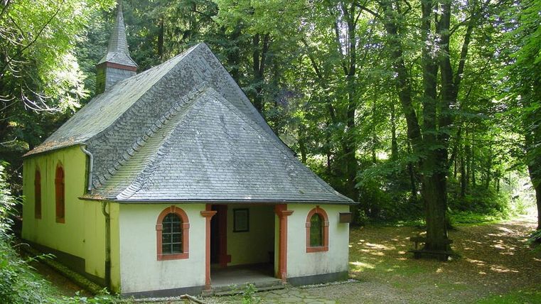 Eine kleine Kirche umgeben von dichtem Wald. Das Gebäude hat ein spitzes Dach und bunte Fenster.