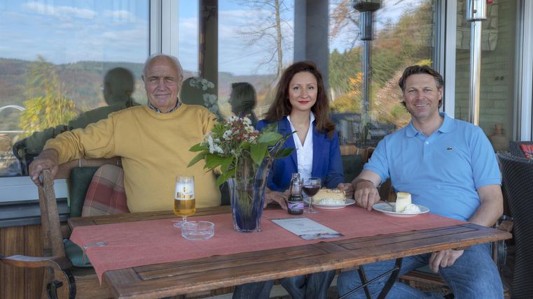 Drei Personen sitzen entspannt an einem Tisch im Freien. Im Vordergrund stehen Getränke und ein Stück Kuchen, mit einer schönen Aussicht im Hintergrund.