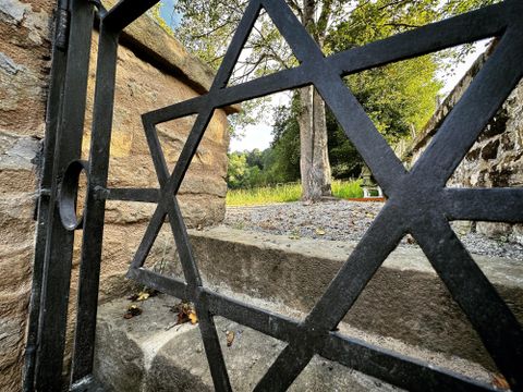 Ein geöffneter schwarzer Metallzaun mit einem Davidsternmuster. Dahinter ist ein grüner Garten mit einem Baum und steinigen Wegen.