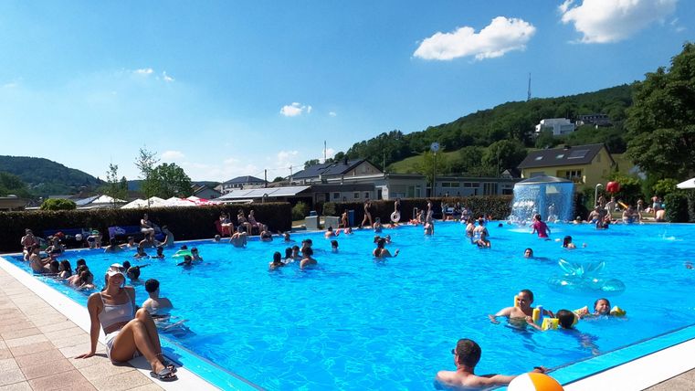 Ein Schwimmbad voller Menschen an einem sonnigen Tag. Im Hintergrund sind grüne Hügel und einige Sonnenschirme zu sehen.