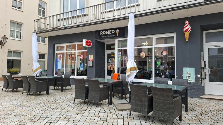 Un café avec des places en extérieur et des parasols. Le bâtiment a de grandes fenêtres et une atmosphère accueillante.