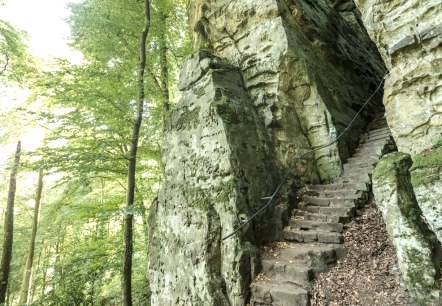 Eine steile Steintreppe f&uuml;hrt durch eine bewaldete Schlucht. Die Felsen sind mit Moos bedeckt, und ein Seil dient als Gel&auml;nder.