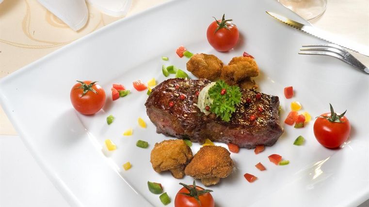 Ein saftiges Steak auf einem weißen Teller, garniert mit frischen Tomaten und buntem Gemüse. Daneben befinden sich knusprige Beilagen.