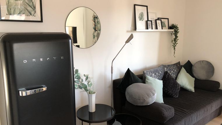 Een moderne woonkamer met een donkere bank en gezellige kussens. Op de achtergrond zijn een koelkast en decoratieve beelden aan de muur te zien.