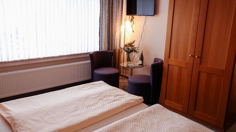 Ein gemütliches Zimmer mit zwei Betten, einem Flachbildfernseher und einer kleinen Sitzecke. Die großen Fenster lassen viel Tageslicht herein.