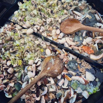 Une grande poêle avec un mélange de fruits de mer frais, de légumes et d'herbes. Deux cuillères en bois sont prêtes à remuer.