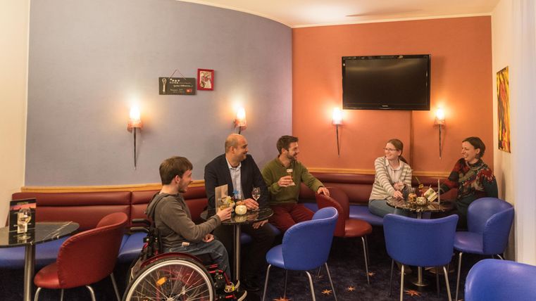 Eine Gruppe von Menschen sitzt in einem gemütlichen Raum und genießt Getränke. Die Sitzgelegenheiten sind bunt, und im Hintergrund ist ein Fernseher zu sehen.