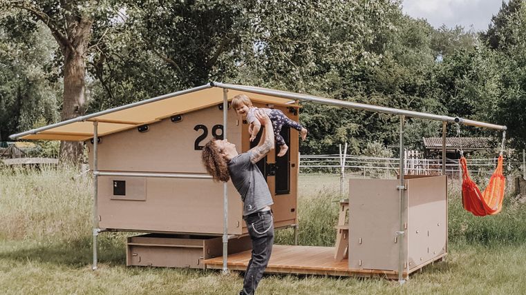 Ein modernes Tiny House steht auf einer grünen Wiese. Ein Erwachsener hebt ein Kind in die Luft, während eine Hängematte daneben hängt.