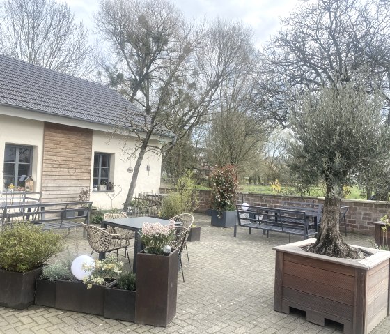 Ein gemütlicher Außenbereich eines Cafés mit Tischen, Stühlen und Pflanzen. Im Hintergrund ein Gebäude mit Holzelementen und Bäumen., © TI Bitburger Land