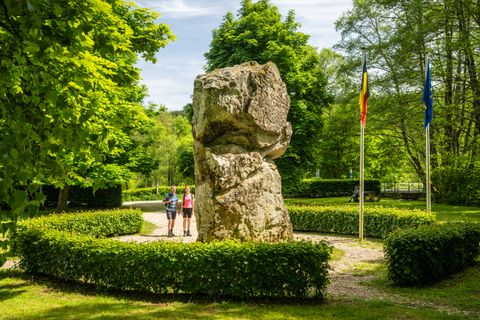 Zwei Personen stehen vor einem großen Felsen im Grünen, flankiert von zwei Flaggen.