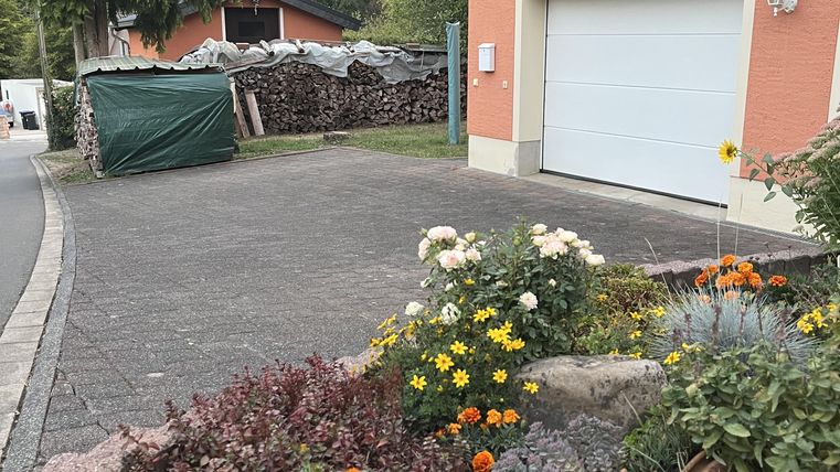 A well-tended garden with colorful flowers and a round decorative ball. In the background, a house and a garage door are visible, surrounded by green trees.