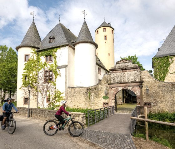 Two cyclists ride past Rittersdorf Castle on the Nims cycle path. The castle has round towers and is overgrown with ivy., &copy; Eifel Tourismus GmbH, Dominik Ketz