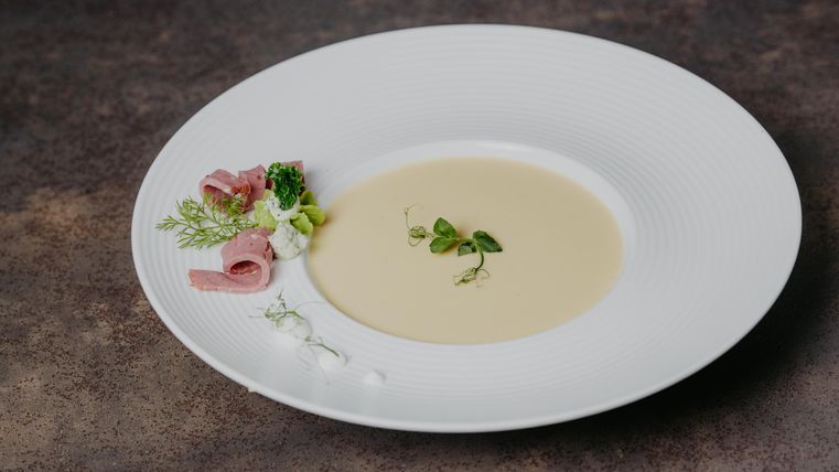 Eine cremige Suppe in einem weißen Teller, elegant angerichtet. Neben der Suppe befinden sich Schinken und frische Kräuter.