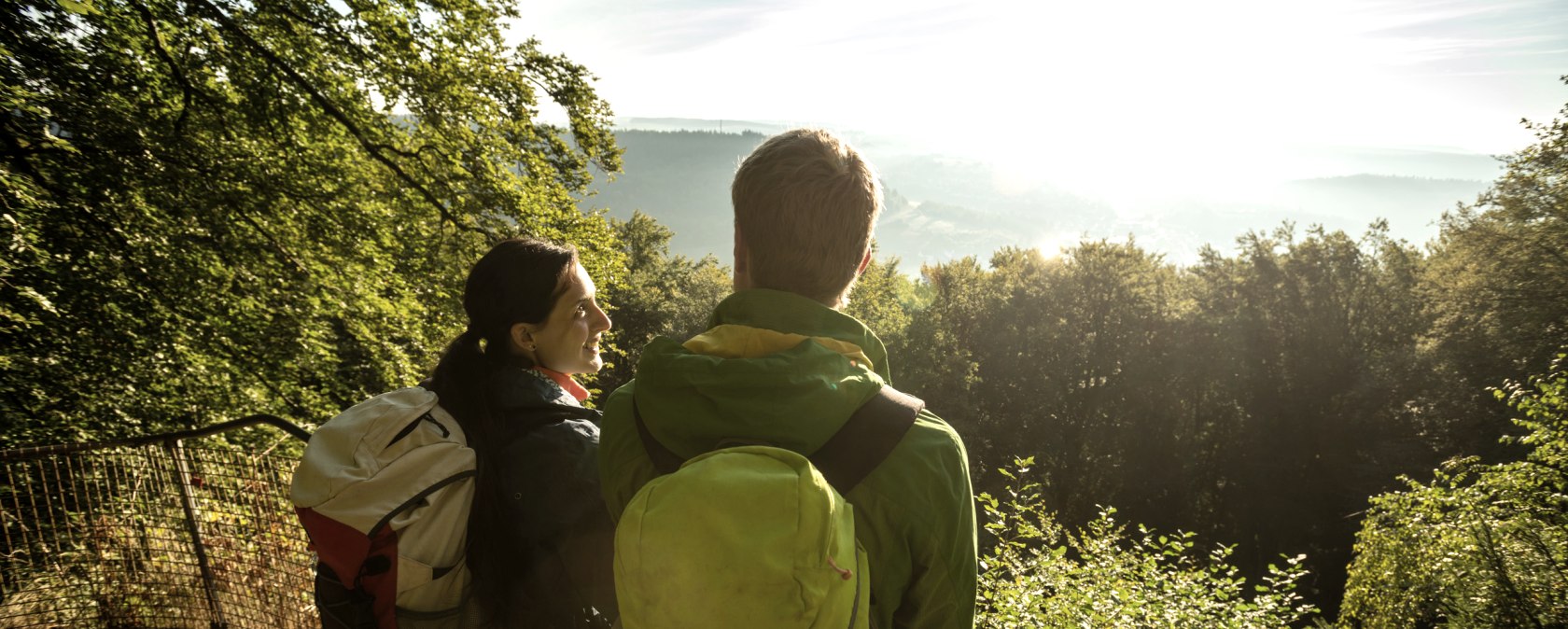 Two hikers with rucksacks stand at a railing and look out over the sunny, wooded landscape., &copy; Dominik Ketz