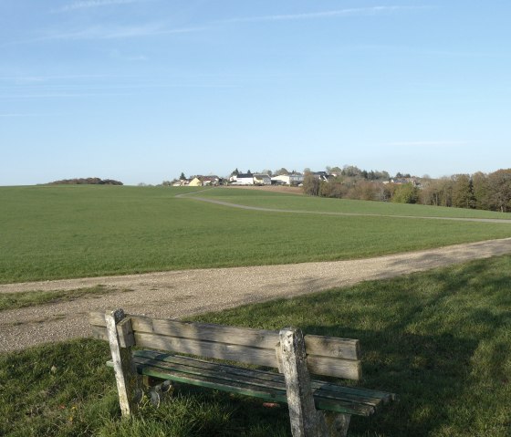 Prairie verte avec banc et chemin, en arrière-plan un village sous un ciel bleu., © Christian Calonec-Rauchfuss, Felsenland Südeifel Tourismus GmbH