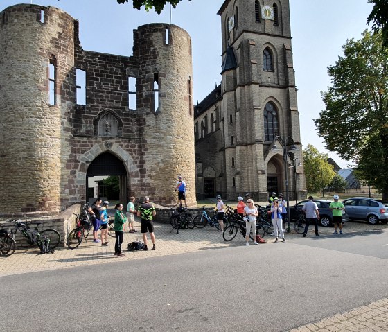 Radfahrergruppe vor einem historischen Tor und einer Kirche, umgeben von Bäumen und geparkten Autos., © Tourist-Information Bitburger Land