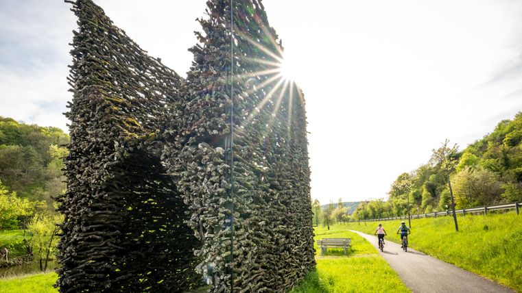 Eine kunstvolle Struktur aus Ästen steht am Wegesrand. Zwei Radfahrer fahren vorbei, während die Sonne aufgeht.