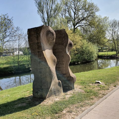 Stenen sculptuur aan de oever van de rivier, omgeven door groen landschap en bomen. Een heldere hemel en een pad zijn ook zichtbaar., © TI Bitburger Land, Steffi Wagner